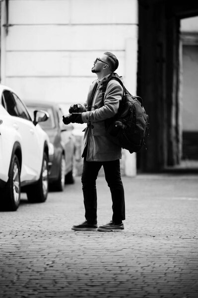 Модный фотограф стоит в очаровательной городской обстановке, запечатлевая моменты с камерой, когда вечерний свет бросает уникальные тени на улицах города.