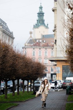 Bir kadın çıplak ağaçlarla çevrili, güzel tarihi binalar ve arka plandaki önemli bir kuleyi gözler önüne seren tuhaf bir caddede canlı bir şekilde yürümekten hoşlanır..