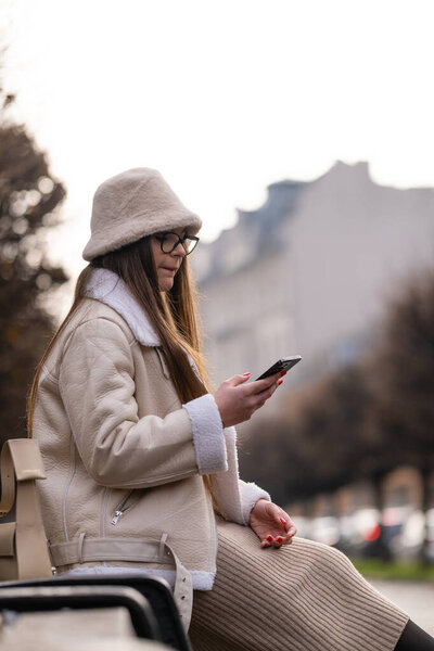 A young woman dressed warmly in a stylish coat and hat sits on a bench, absorbed in her phone on a chilly afternoon, surrounded by leafless trees and distant buildings.