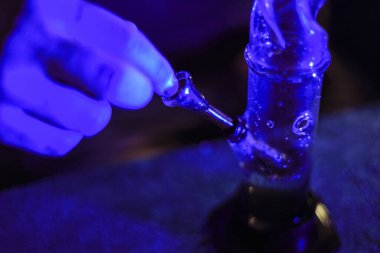 Erkek eli marihuanayla nargileyi tıkar. Uyuşturucu ve uyuşturucu bağımlısı. Adam ot içiyor. Neon mavi ışık. CBD veya THC