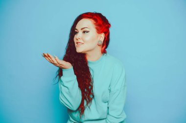 Mavi arka planda mavi kazaklı bir kadın poz veriyor. Uzun saçları ve kulaklarında tünelleri olan güzel bir esmer. Göz kalemi ve moda makyaj. İtaatkar öpücük.