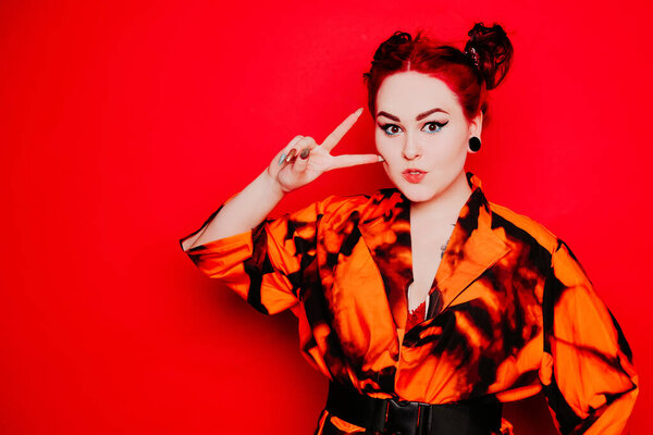 Beautiful girl on a red background. Red hair and tunnels in the ears, red dress and lips. Anime poses. The girl with eyeliner and two buns on her head grimaces, smiles, has fun. Peace gesture