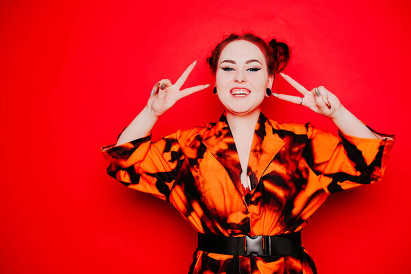 Beautiful girl on a red background. Red hair and tunnels in the ears, red dress and lips. Anime poses. The girl with eyeliner and two buns on her head grimaces, smiles, has fun. Peace gesture