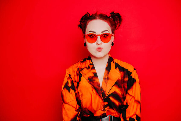 Cute girl on a red background. Red hair and tunnels in the ears, red dress and lips. Anime poses. A girl in orange sunglasses and two buns on her head looks at the camera.