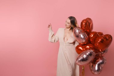 Woman with red balloons. Joyful model having fun, playing and celebrating Valentine's day in long pink dress pointing to empty seat. Surprised, smiling, discounts, product