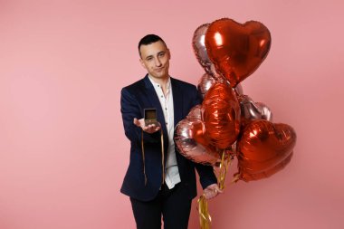 A man with heart balloons on a pink background holds out a box with a wedding ring. Betrothed on Valentine's Day, man proposing, sad face, unhappy, bad sad.