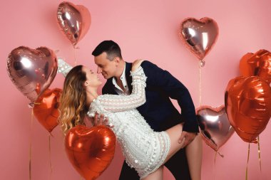 A couple in evening dresses are kissing while dancing against a background of red and pink balloons in the shape of a heart on a pink background. Vespers on Valentine's Day or March 8, Women's Day
