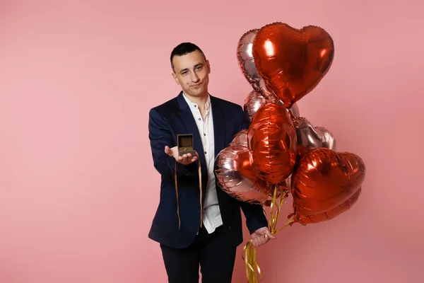 A man with heart balloons on a pink background holds out a box with a wedding ring. Betrothed on Valentine's Day, man proposing, sad face, unhappy, bad sad.
