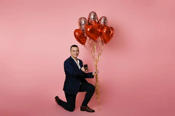 A man in a suit with red balloons on a pink background holds out a box with a wedding ring. Engaged on Valentine\'s Day, the man proposes, smiles