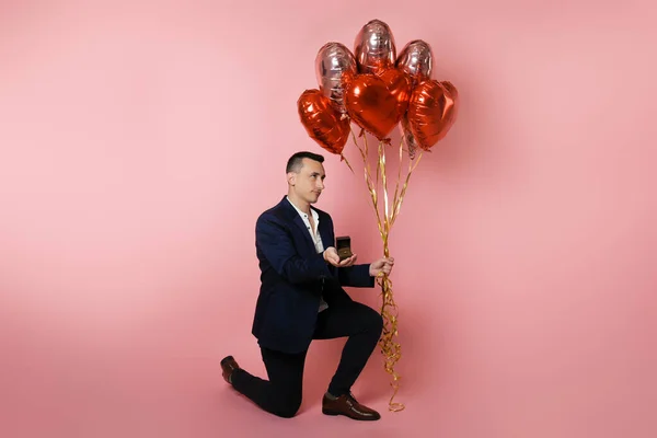 A man with heart balloons on a pink background holds out a box with a wedding ring. Betrothed on Valentine\'s Day, man proposing, sad face, unhappy, bad sad.