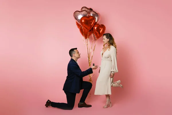 A man with heart balloons on a pink background holds out a box with a wedding ring to a girl. Engaged on Valentine's Day, man proposes, Valentine's Day, February 14, love