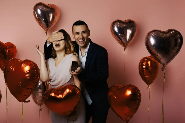 A man with heart balloons on a pink background holds out a box with a wedding ring to a girl. Engaged on Valentine\'s Day, man proposes, Valentine\'s Day, February 14, love