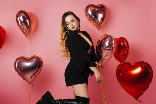 Gorgeous girl in a black dress and corset, in jewelry and raincoats with red balloons on a pink background. Fashion and fashion, Valentine\'s Day