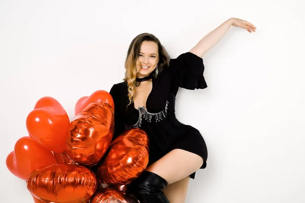 Sexy woman in a black dress and corset, in jewelry and boots in red balloons on a white background. Fashion and fashion, Valentine's Day, dancing, party