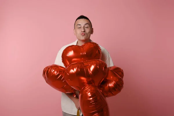 A man is holding red balloons in the shape of a heart on a pink background. Valentine\'s Day, love, February 14. Sends an air kiss to the camera