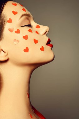Yüzünde kalp çıkartması olan bir kadının portresi. Artistik profesyonel makyajın moda fotoğrafı, gri arka planda kırmızı dudaklı bir kadın. Aşk ve duygular, tarz 14 Şubat, Sevgililer Günü