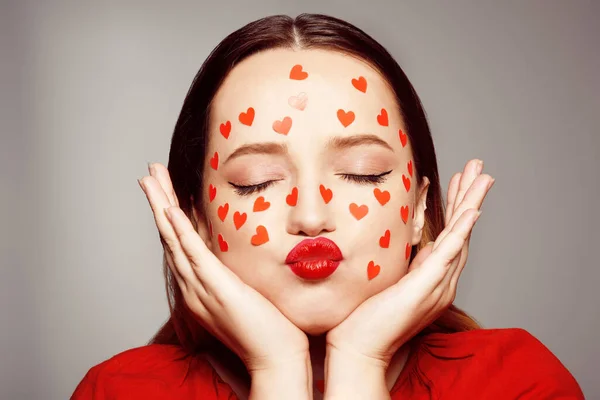 Portrait of a woman with stickers of hearts on her face. The girl blows an air kiss on the camera, on a gray background. Self-love, skin care, makeup, courses for women, dating