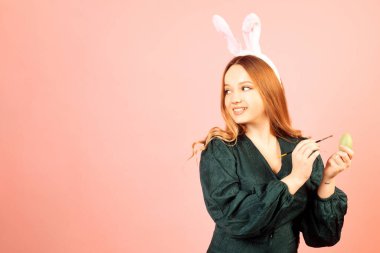 Tavşan kulaklı güzel bir kız pembe arka planda paskalya yumurtaları boyuyor. Gülümseyen ve gülen bir kadın, yeşil elbiseli güzel saçlı bir model..