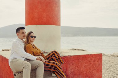 Sahildeki deniz fenerinin yanında, denizin ve dağların arkasında bir çift oturuyor. Tatil aşkı, balayı, seyahat. şık bir çift.