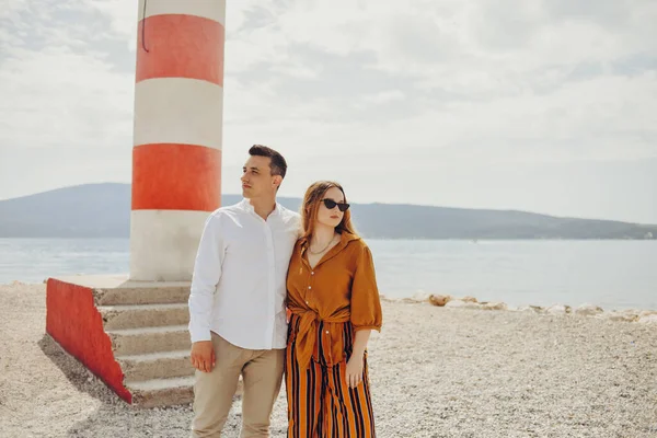 夫婦は海と山を背景にビーチの灯台の近くに立っています 休暇の愛 新婚旅行 スタイリッシュなカップル笑いと笑顔 — ストック写真
