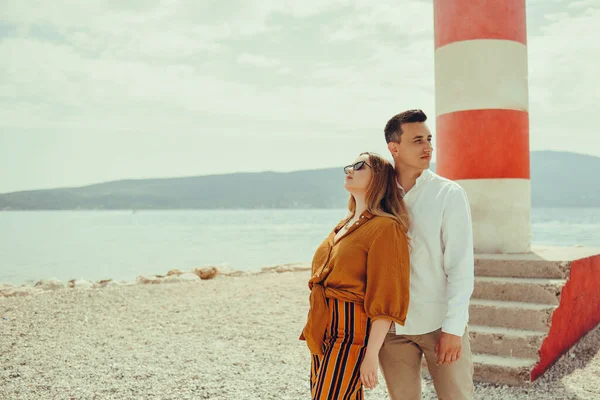 夫婦は海と山を背景にビーチの灯台の近くに立っています 休暇の愛 新婚旅行 スタイリッシュなカップル笑いと笑顔 — ストック写真