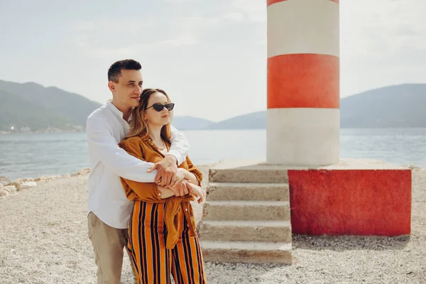 一对夫妇站在海滩上的一座灯塔旁边 面对着大海和群山 度假爱 度蜜月 年轻貌美的夫妻笑着 — 图库照片
