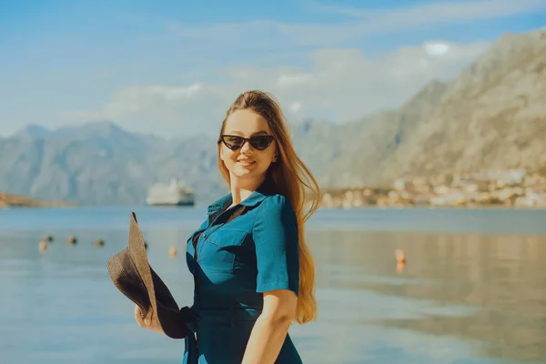 A woman in sun glasses and a hat stands against the backdrop of the sea and mountains. Vacation and outdoor recreation. Girl with long hair on vacation