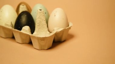 Paskalya geliyor, çıplak arka planda boyalı yumurtalarla dolu bir tepsi. Kutlamalar ve gelenekler. Mavi, altın ve beyaz, siyah yumurtalı, tavuk tüyleri uçuşuyor. Plastik yumurtalar, metin için yer.