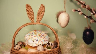 Easter holidays, cute basket with rabbit ears on a green background. Easter cakes, colored eggs, and decorations on Pussy willow. Traditions and food, Easter bread, moss and chicken feathers, hay.