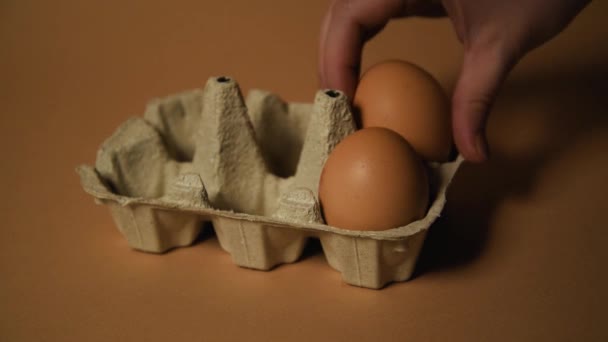 ヌードブラウンの背景にトレイの卵 女性の手で一つの卵をトレイに入れます レシピ エコ製品と食品 トレイに6個の卵 品質の製品 — ストック動画