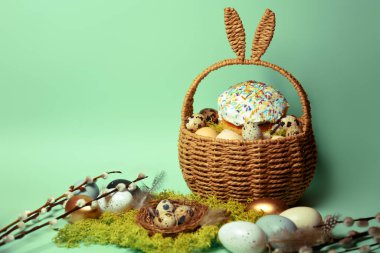 Easter decorations in a basket with rabbit ears. Easter cake and eggs on moss, quail eggs and feathers on mint green background. pussy willow Place for text