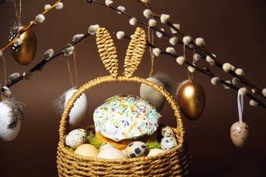 Easter holidays, cute basket with bunny ears on brown background. Easter cakes with colored sprinkles, colored golden eggs, and pussy willow decorations. Nest with eggs, Traditions