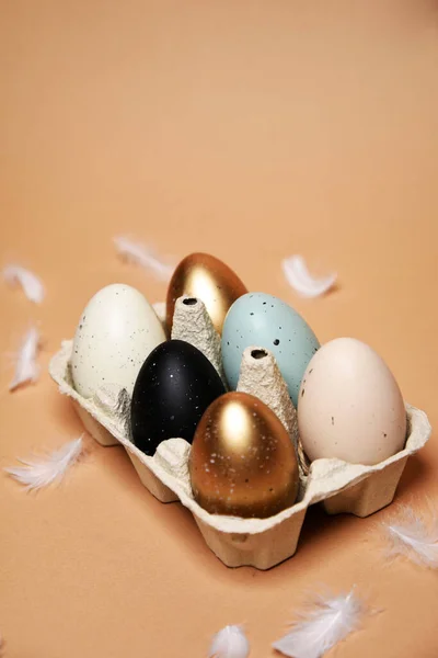Easter is coming, a tray with painted eggs, on a nude background. Celebrations and traditions. Blue, gold and white with black egg, chicken feathers flying. Plastic eggs, place for text.
