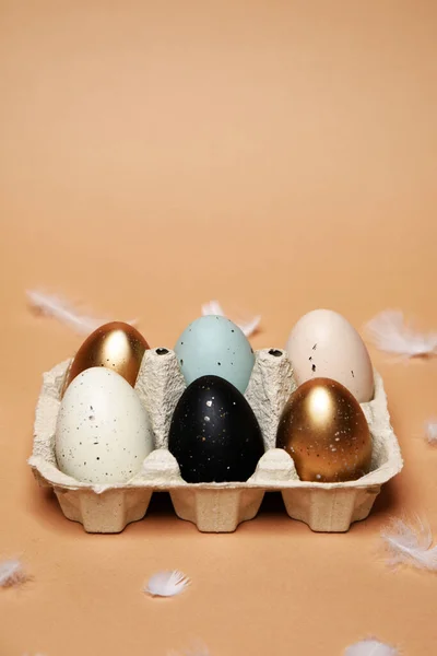 Easter is coming, a tray with painted eggs, on a nude background. Celebrations and traditions. Blue, gold and white with black egg, chicken feathers flying. Plastic eggs, place for text.