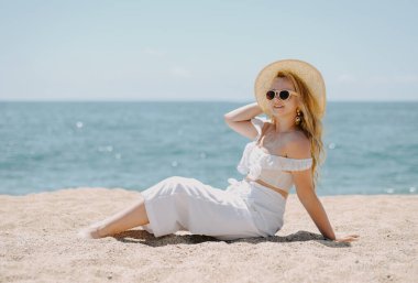 Beyaz elbiseli şık, uzun bacaklı bir model okyanusun arka planında sahilde oturuyor ve hayatın tadını çıkarıyor. Tatil, tatil, yaz havası. Güzel mücevherler içinde sarışın, zengin bir kadın..