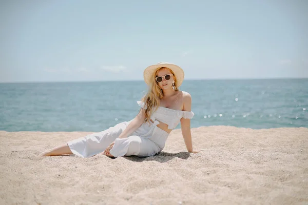 A chic fashionable long-legged model in a white outfit sits on the beach against the backdrop of the ocean and enjoys life. Vacation, vacation, summer mood. Blonde in beautiful jewelry, rich woman.