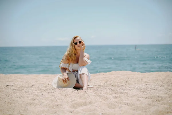 白い服を着たシックなファッショナブルなロングレッグモデルが海を背景にビーチに座り 人生を楽しんでいます 夏の気分 美しいジュエリーでブロンド 豊かな女性 — ストック写真