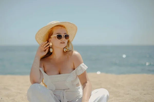 白い服を着た幸せな女の子は 海や海のビーチを背景に砂の上に座っています 女性は笑顔と笑い 休暇と喜び ファッションモデル 美しいジュエリー イヤリング サングラス — ストック写真