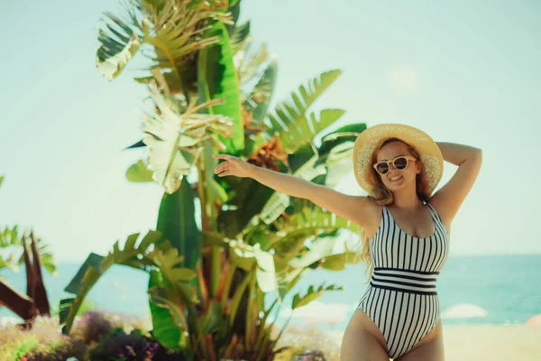 一个金发碧眼的夏天在海滩上摆姿势 一个戴着帽子和时髦太阳镜的女人站在沙滩上 海洋或海洋为背景 在棕榈树下休息 — 图库照片