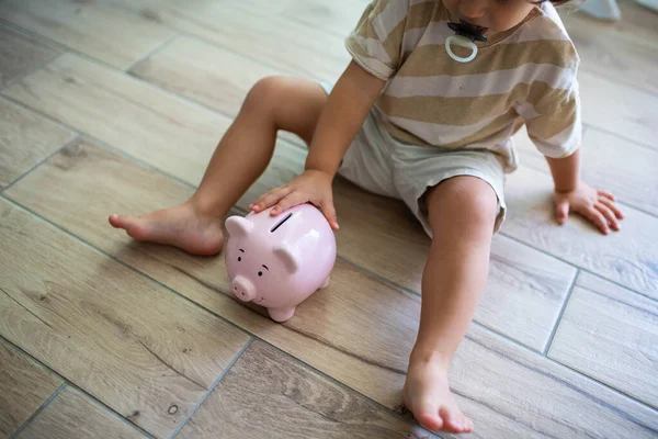 白种人蹒跚学步 带着抚慰器 坐在一个笑着的小猪银行旁边的地板上 为孩子省钱的概念 — 图库照片#