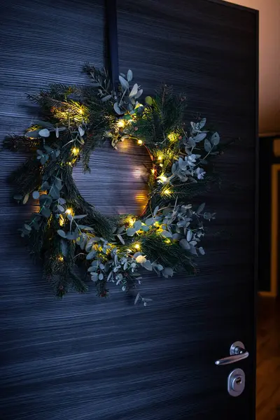 Foraged diy pine and eucalyptus Christmas wreath, with illuminated string lights, hanging on opening door. Vertical shot.