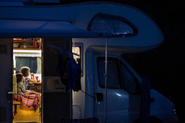 Two kids, not looking, sitting in a camper van at night, seen from outside through the open door. clipart