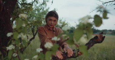 Romantik kadın, kahverengi gömlekli güzel kadın model ağaçların dallarında dikilip kameraya bakıyor.