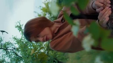 Güzel romantik bir kadının dikey video portresi, güzel sarışın manken kamera önünde hareket ediyor, elma ağacının dallarında gülümsüyor.