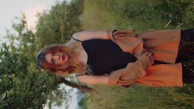 Dikey video romantik kadın, kahverengi gömlekli güzel sarışın manken, elma bahçesindeki bir çayırda poz veriyor.