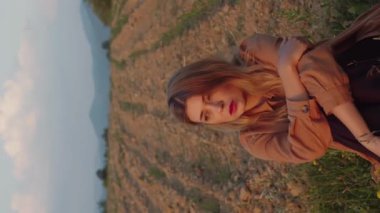 Dikey video oldukça romantik beyaz kadın, güzel sarışın kadın çayırda oturuyor ve batan güneşin altında tarlayı sürüyor.