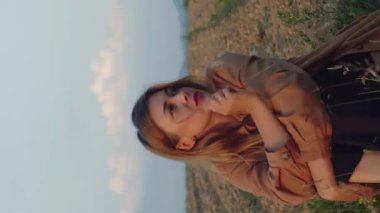 Dikey video romantik beyaz kadın çayırda oturuyor ve batan güneşin ışınlarında tarlayı sürüyor.