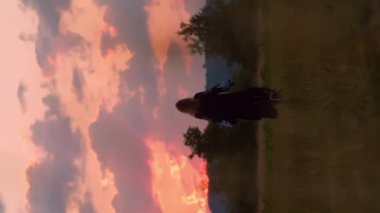 Kırmızı, pembe, sarı gün batımında veya kırmızı gündoğumunda ulusal giyinmiş romantik sarışın kızın dikey video silueti. Köy kavramı