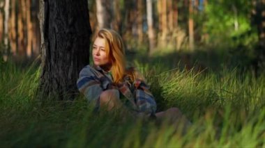 Huzurlu kız, uzun, kalın çimenlerde bir ağacın altında dinleniyor ve rahatlıyor. Parkta rahatlama ve meditasyon kavramında çimlerin üzerinde oturuyor.
