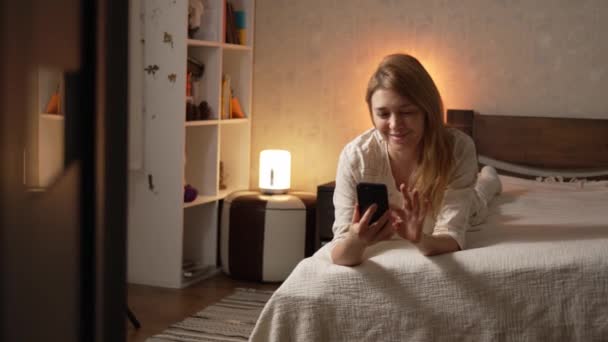 穿着睡衣的年轻而快乐的金发女人躺在床上舒舒服服地在家里聊天 发短信 用智能手机 电话传达周末快乐的微笑 — 图库视频影像